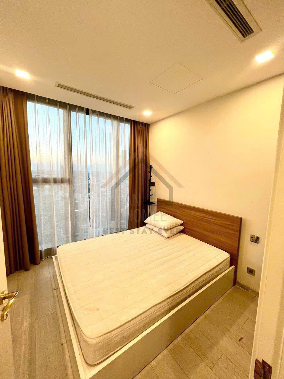 Phòng ngủ nhỏ với giường gỗ hiện đại, nệm trắng và cửa sổ lớn rèm nâu nhìn ra thành phố