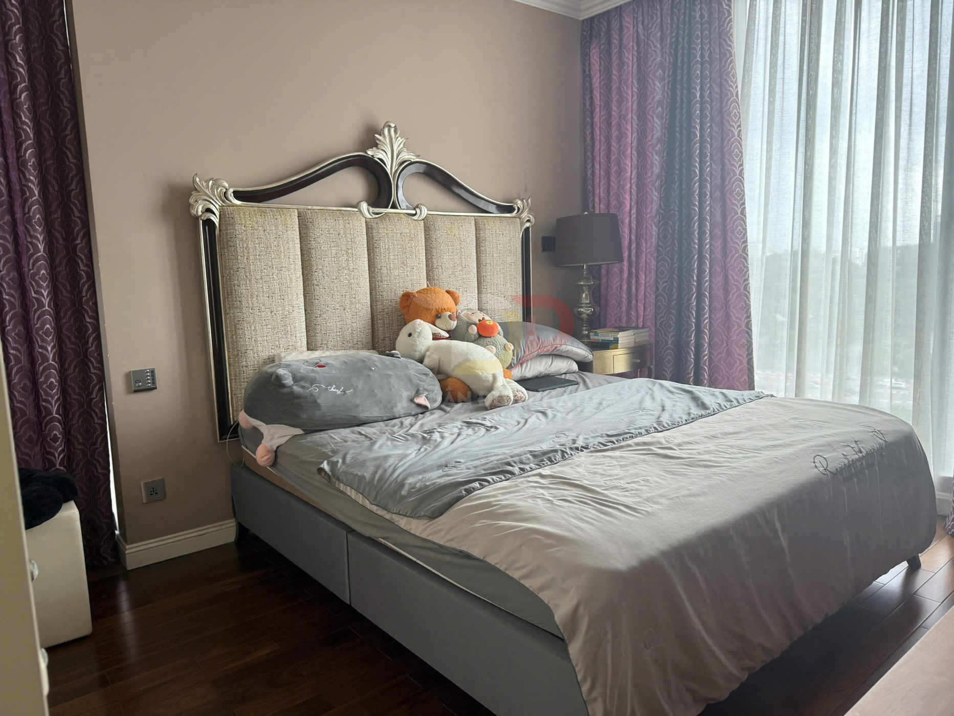 Giường ngủ bọc nệm với chăn ga màu xám và thú nhồi bông, cạnh cửa sổ lớn có rèm tím