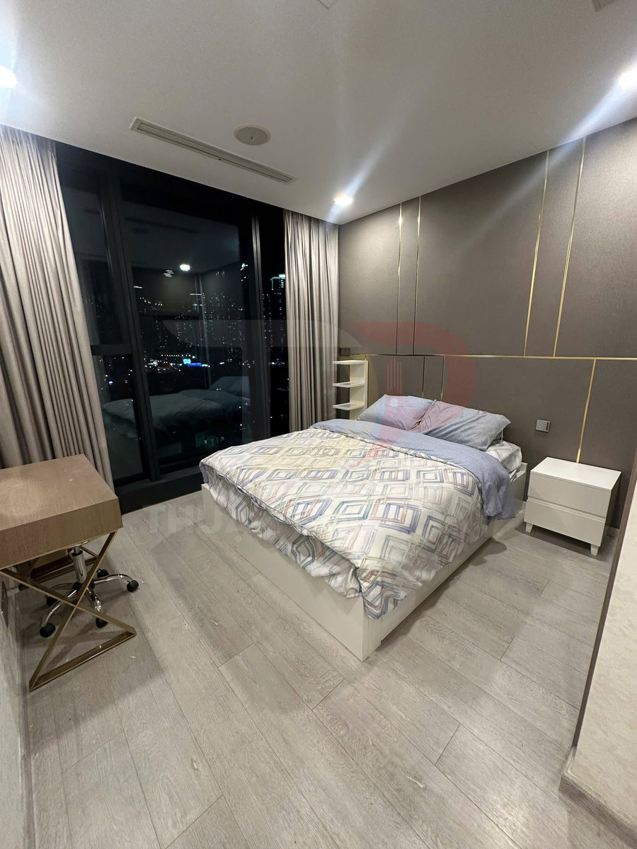 Phòng ngủ hiện đại với giường đôi, ga họa tiết hình học, bàn làm việc nhỏ và cửa kính lớn nhìn ra thành phố ban đêm