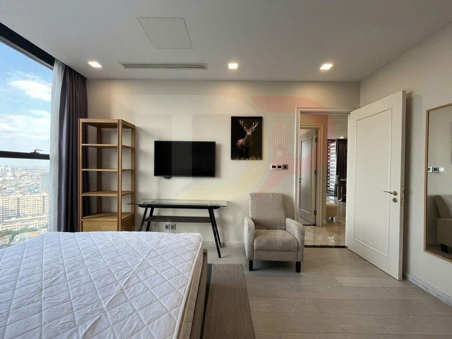 Phòng ngủ chung cư hiện đại với giường nệm trắng, tivi treo tường, kệ gỗ và ghế bành cạnh cửa sổ lớn view thành phố