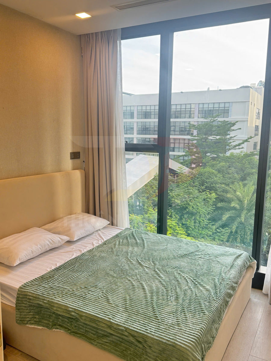 Giường ngủ đôi với chăn xanh và hai gối trắng cạnh cửa sổ kính lớn nhìn ra cây xanh