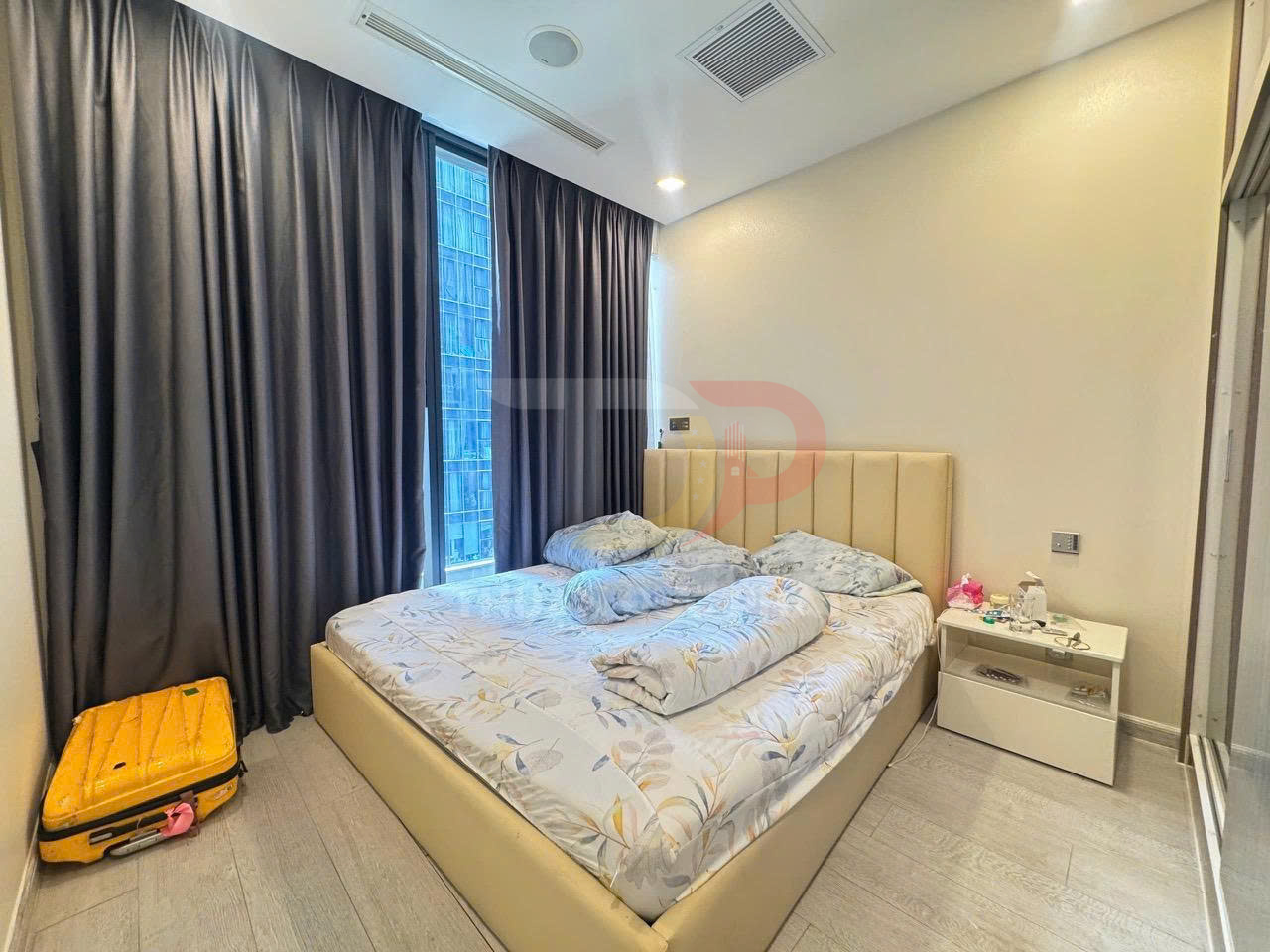 Phòng ngủ nhỏ với giường đôi bọc da màu kem, chăn ga hoa nhã và tủ đầu giường trắng cạnh cửa sổ lớn