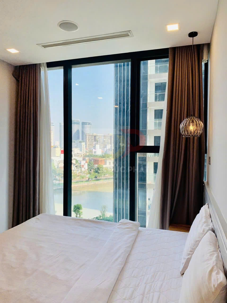 Phòng ngủ chung cư với giường đôi trắng cạnh cửa kính lớn nhìn ra sông và tòa nhà cao tầng