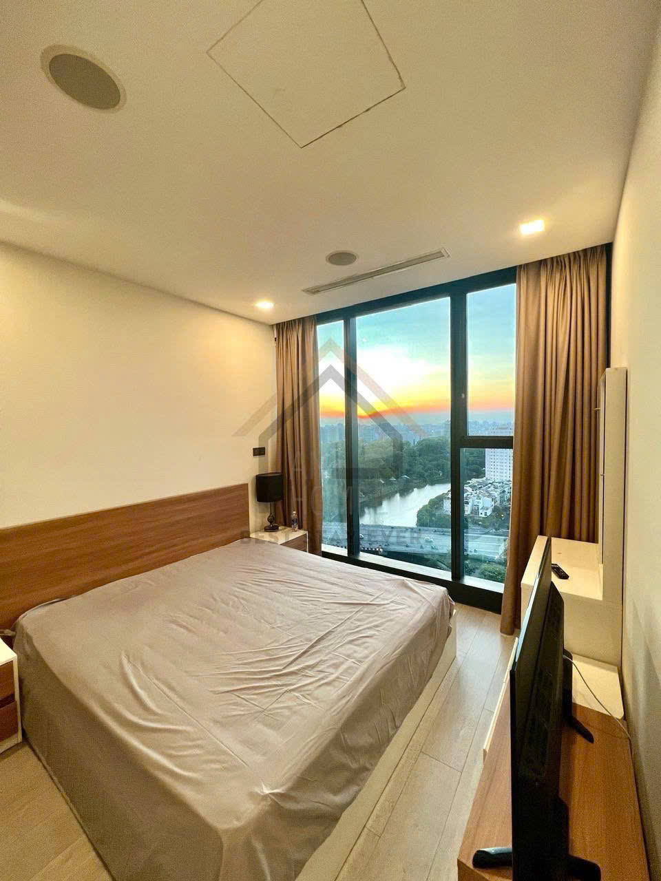 Phòng ngủ chung cư hiện đại với giường đôi, tivi và cửa kính lớn nhìn ra sông lúc hoàng hôn