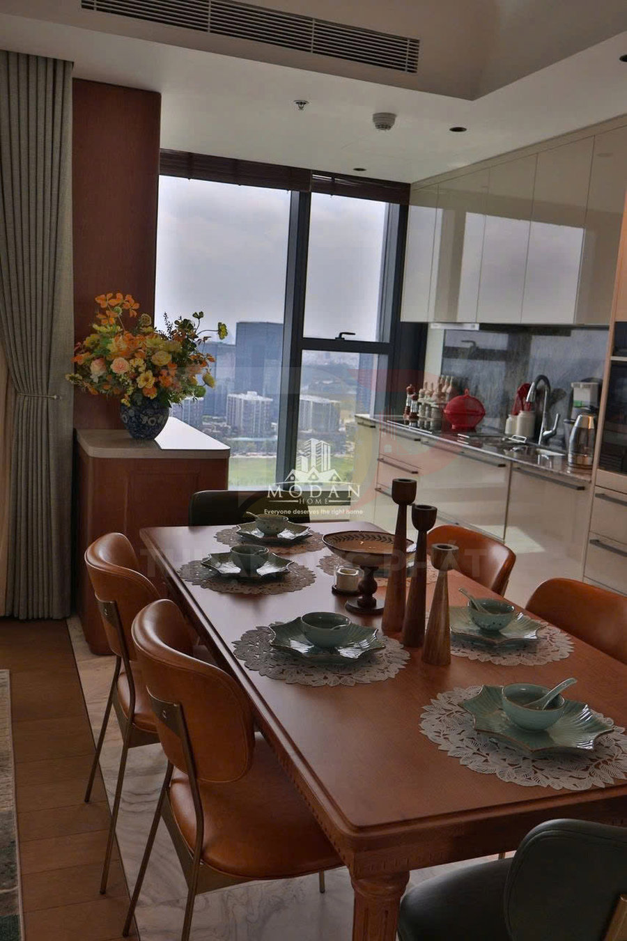 Bộ bàn ăn gỗ bày bát đĩa sứ xanh trong căn bếp chung cư hiện đại có cửa sổ lớn view thành phố