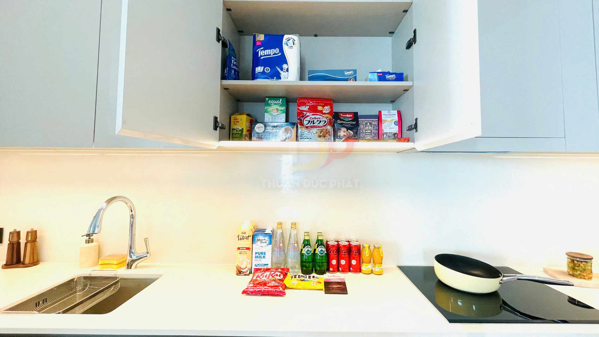 Quầy bếp gọn gàng với tủ đựng đồ ăn vặt, nước ngọt Coca Cola, nước khoáng Perrier và chảo chống dính trên bếp điện