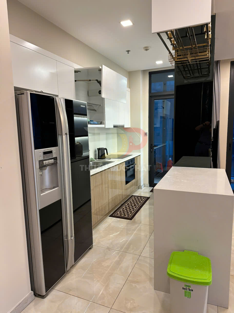 Tủ bếp hiện đại với tủ lạnh side by side, bếp âm, kệ bếp gỗ và thùng rác nhựa nắp xanh