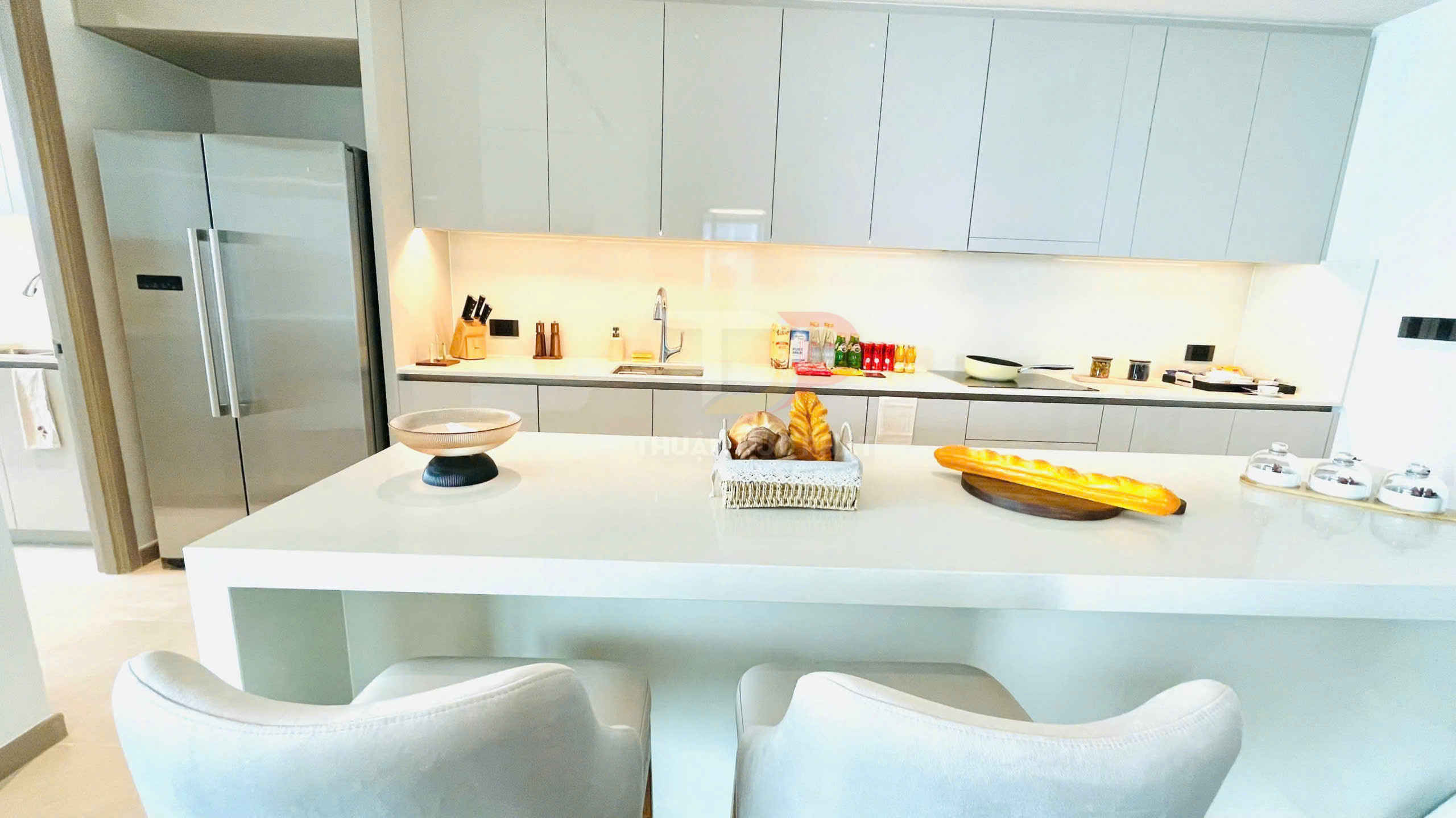 Bếp hiện đại với tủ bếp trắng, đảo bếp lớn, tủ lạnh đôi inox và bánh mì trưng bày trên mặt bàn