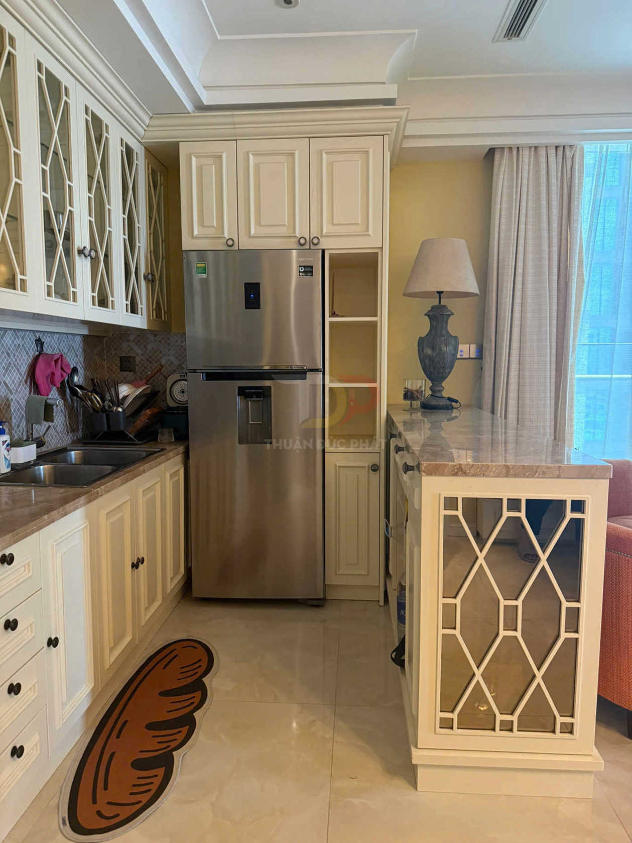 Tủ bếp gỗ màu kem với tủ lạnh inox hai cánh và thảm lót sàn hình ổ bánh mì trong gian bếp hiện đại