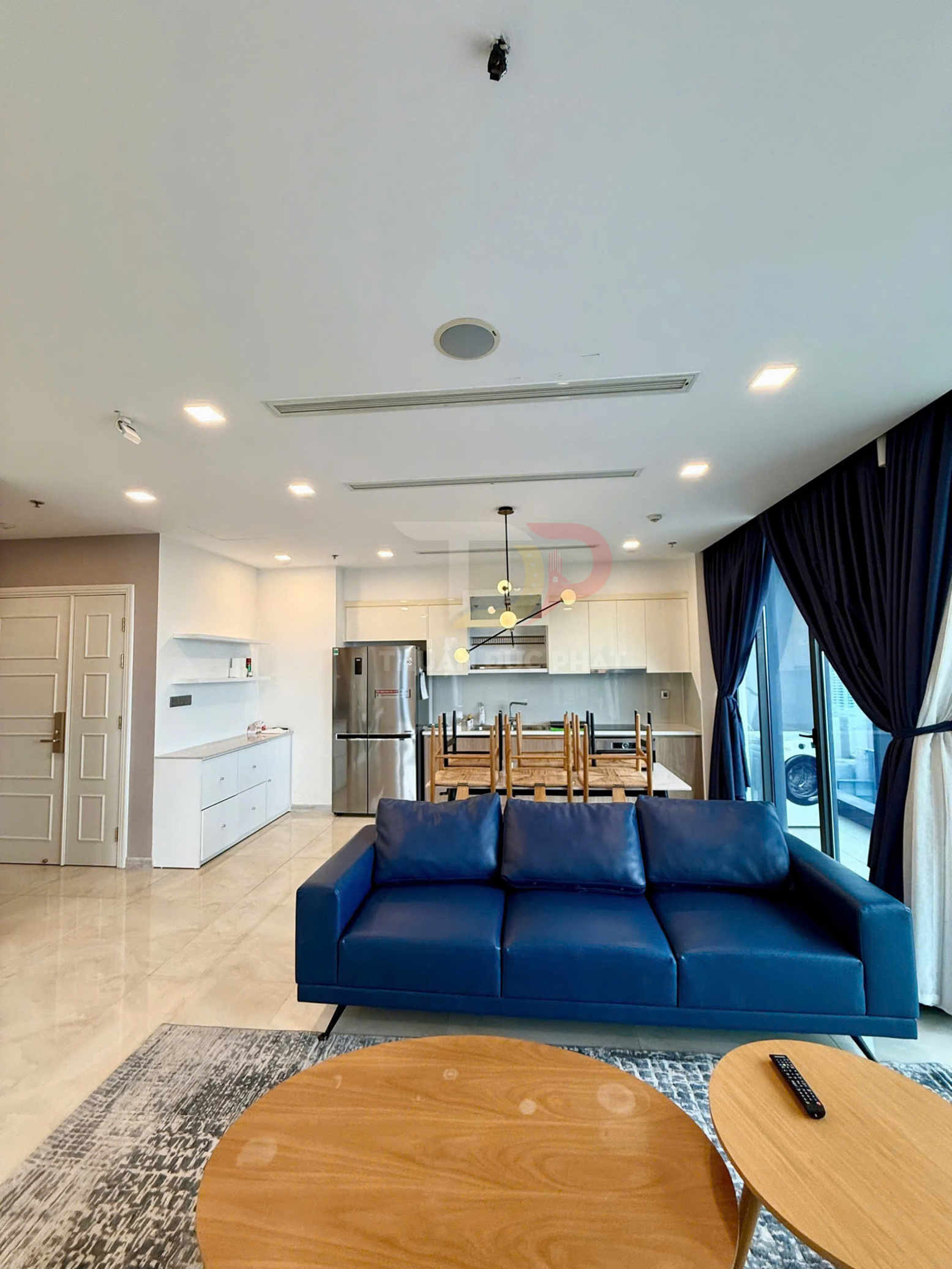 Sofa da màu xanh dương trong phòng khách hiện đại liền bếp với bàn gỗ tròn và rèm cửa tối màu