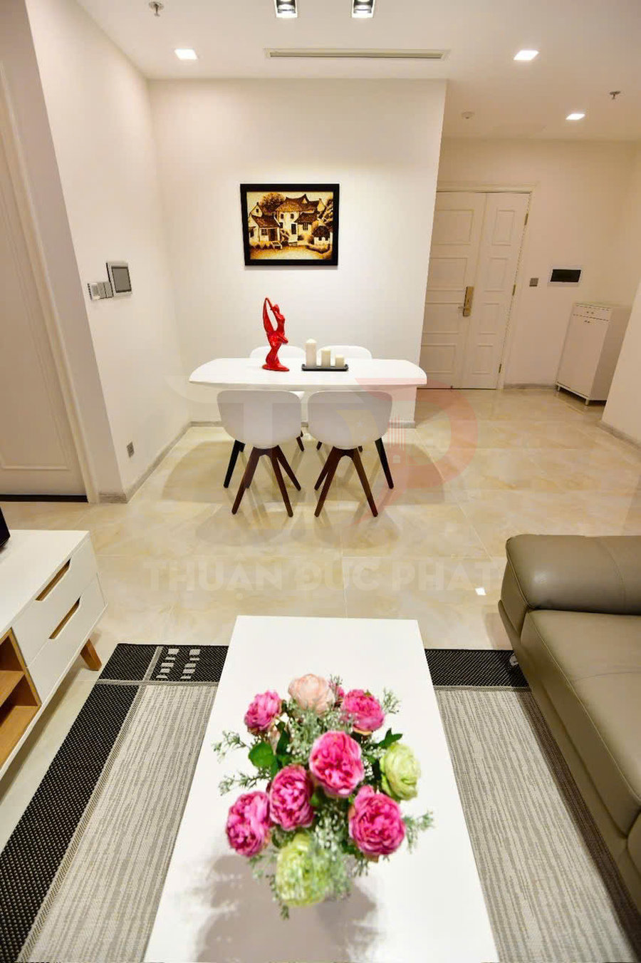 Bàn trà phòng khách với bình hoa hồng trang trí, phía sau là bàn ăn trắng và ghế hiện đại trong căn hộ sáng sủa