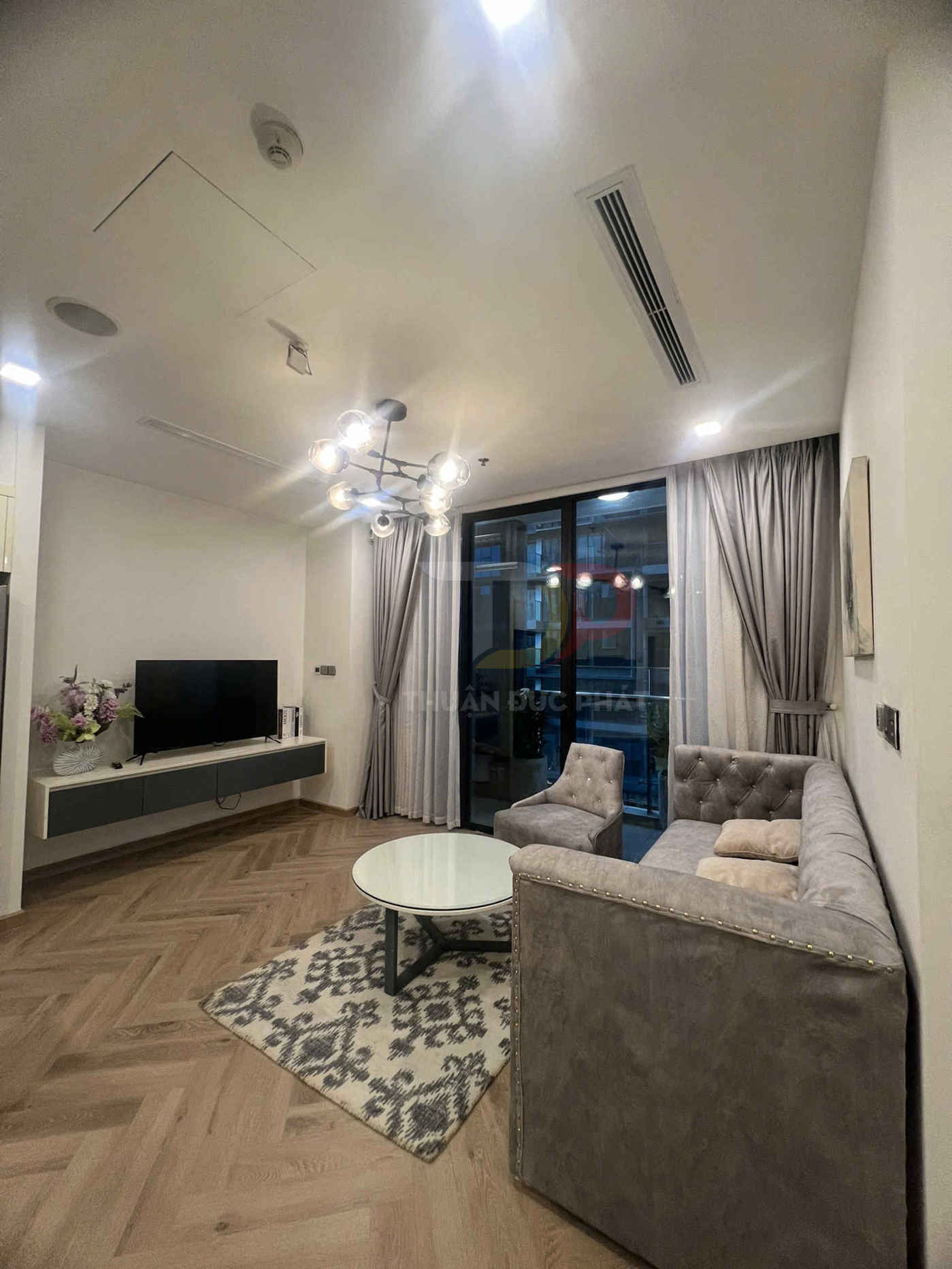 Phòng khách chung cư hiện đại với sofa bọc nỉ xám, bàn trà tròn trắng và kệ tivi treo tường cạnh cửa kính lớn