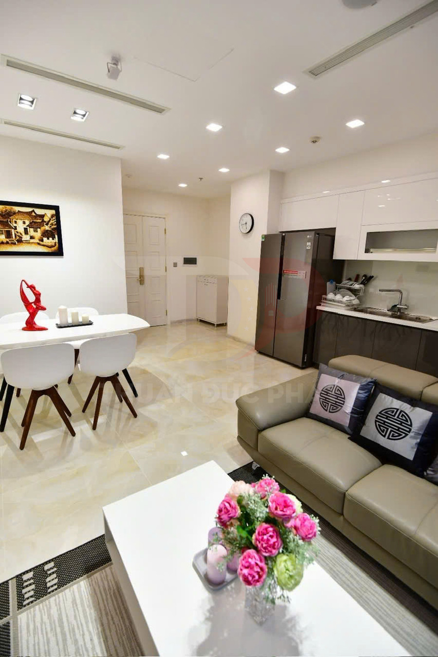 Phòng khách và bếp chung cư hiện đại với sofa da, bàn trà hoa tươi, bàn ăn trắng và tủ bếp gọn gàng