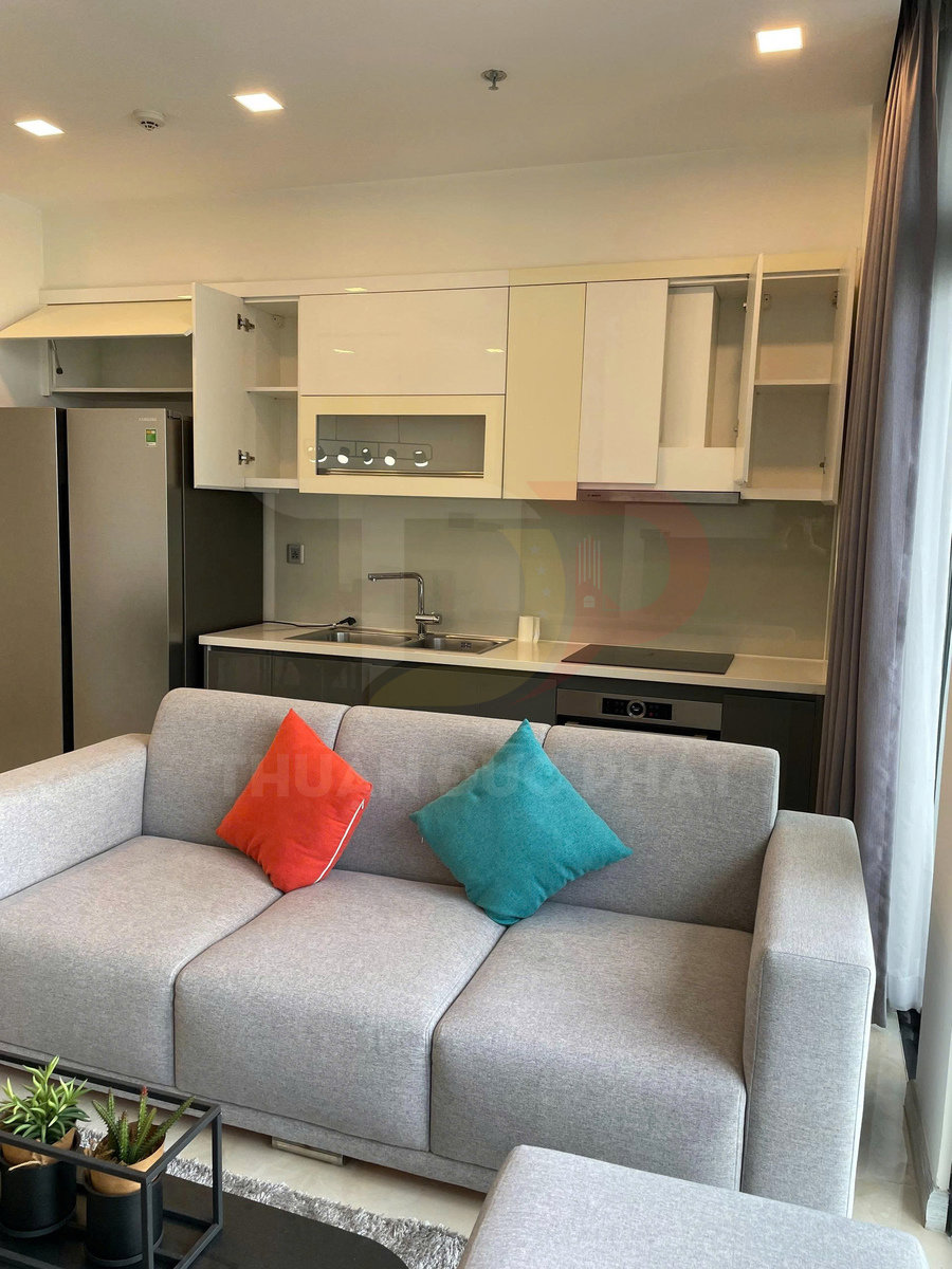 Sofa vải xám với gối tựa màu cam và xanh đặt trong phòng khách liền bếp hiện đại có tủ bếp và tủ lạnh