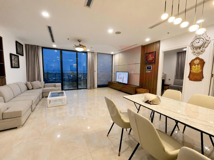 Phòng khách chung cư hiện đại với sofa chữ L màu be, bàn ăn mặt đá và cửa kính lớn nhìn ra ban công