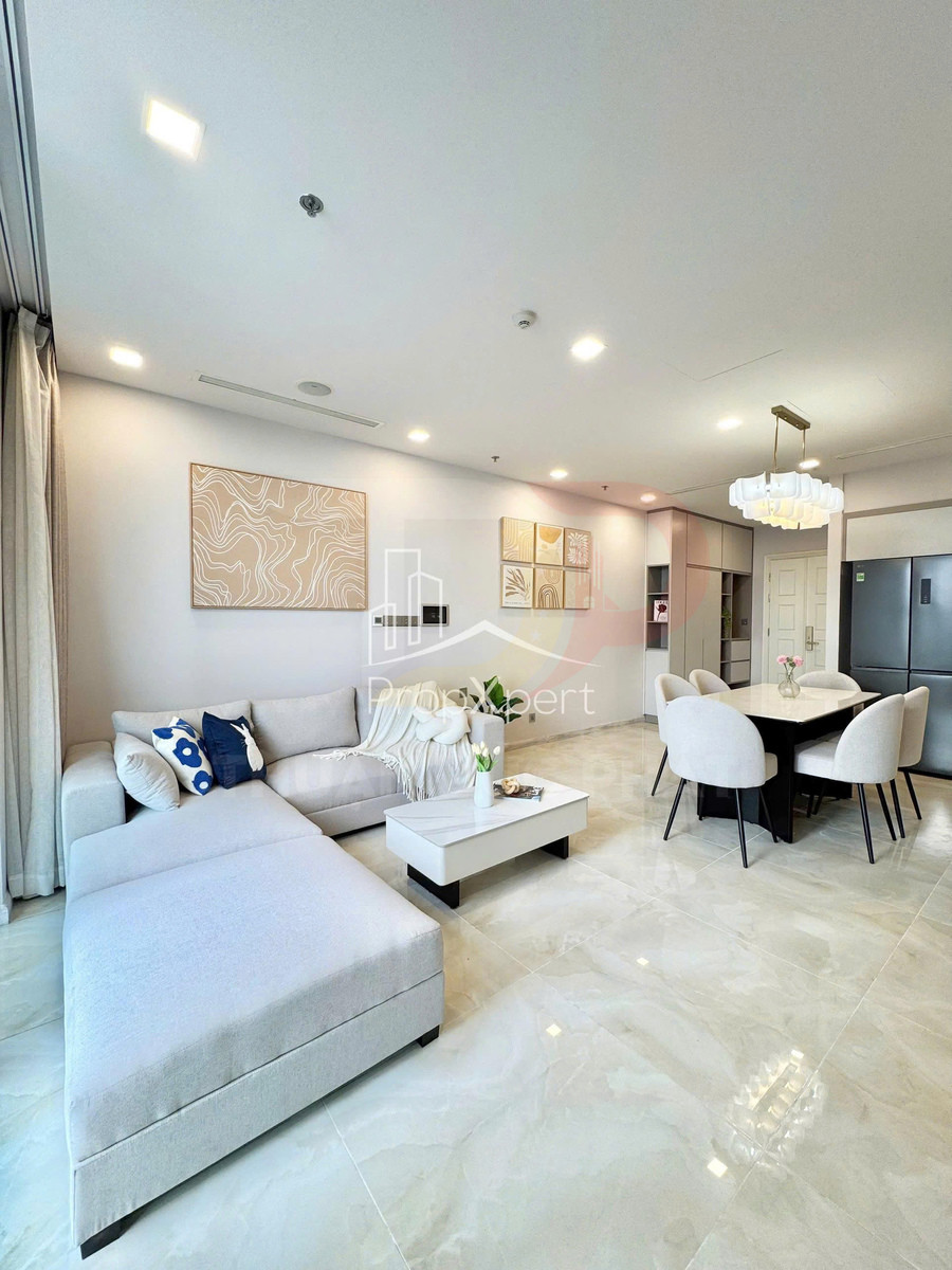 Phòng khách căn hộ hiện đại với sofa góc màu xám nhạt, bàn trà thấp và bộ bàn ăn 6 ghế cạnh bếp