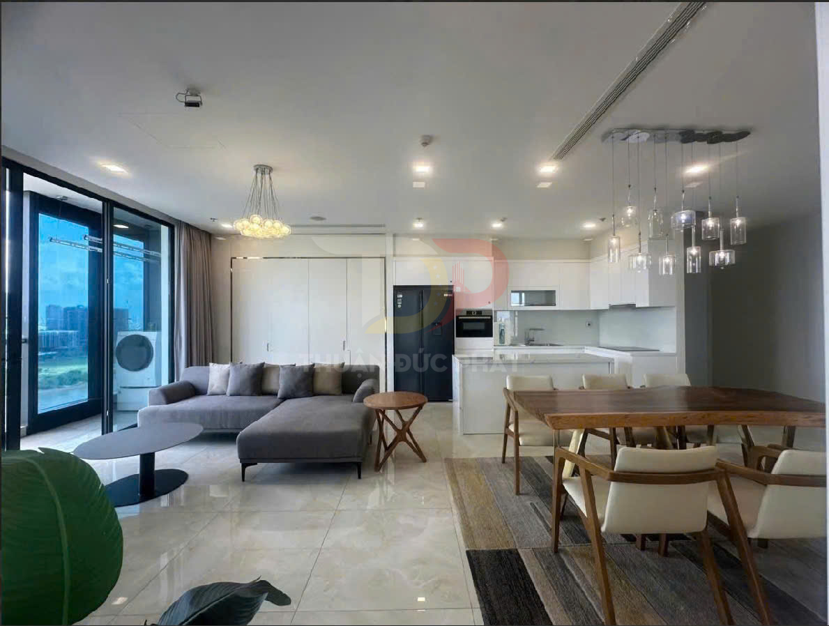 Phòng khách và bếp căn hộ hiện đại với sofa góc xám, bàn ăn gỗ và tủ bếp trắng liền kề ban công kính