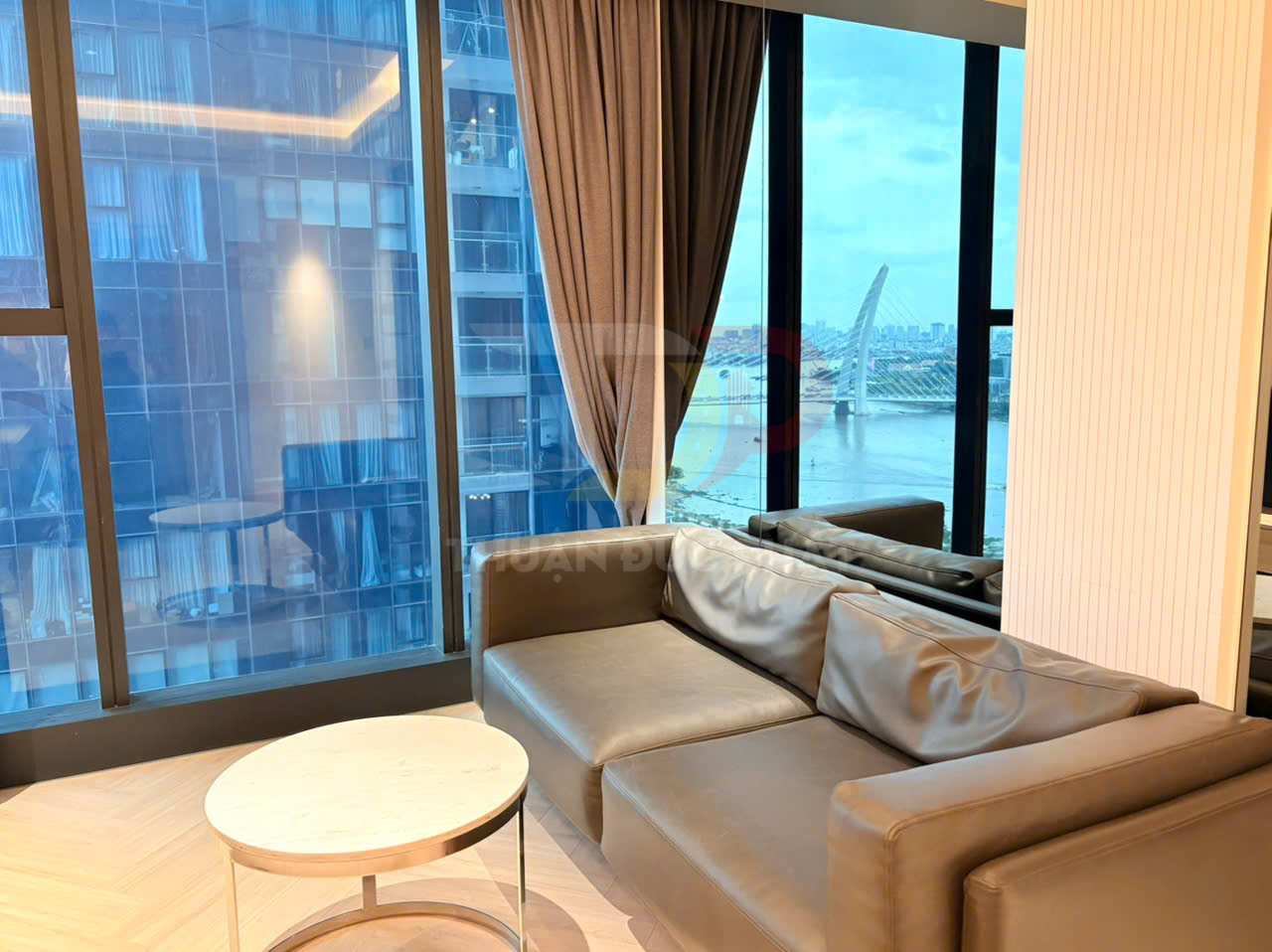 Sofa da góc màu nâu đặt cạnh bàn trà tròn trong phòng khách chung cư view sông và thành phố qua cửa kính lớn