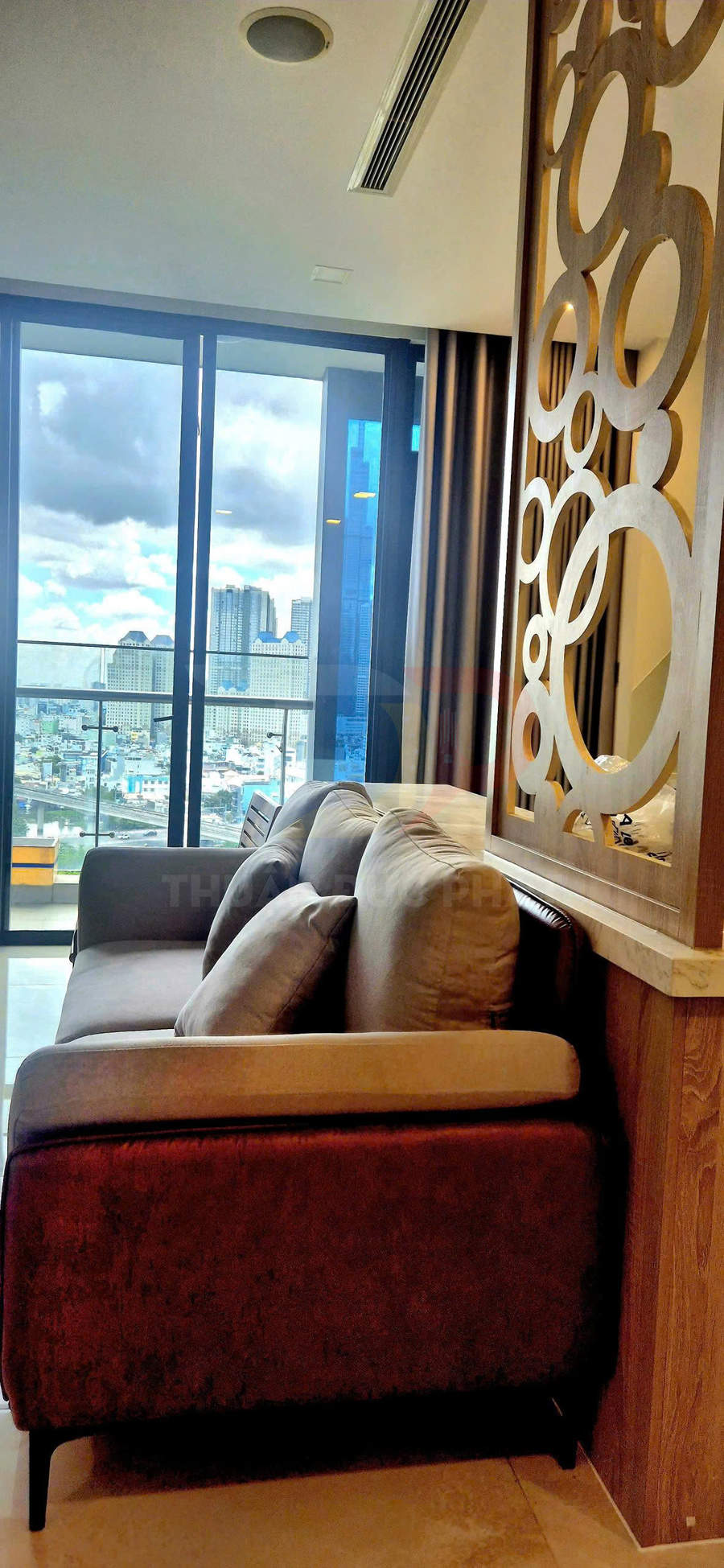 Sofa nỉ màu nâu trong phòng khách chung cư, cạnh vách gỗ trang trí và cửa kính lớn nhìn ra thành phố