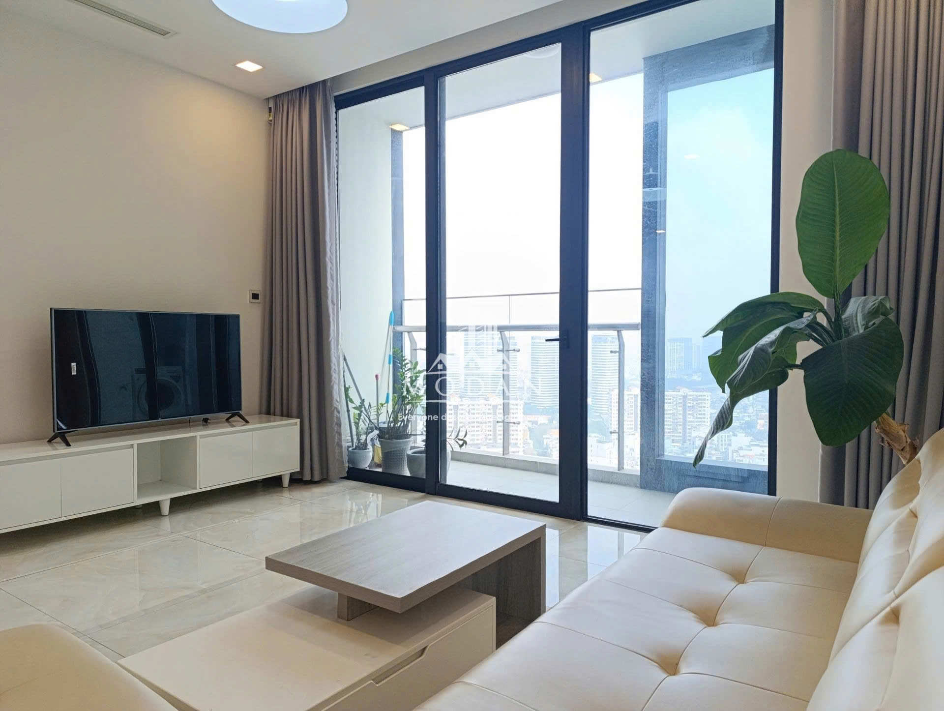 Phòng khách chung cư hiện đại với sofa da màu kem, bàn trà gỗ, kệ tivi trắng và cửa kính lớn nhìn ra ban công