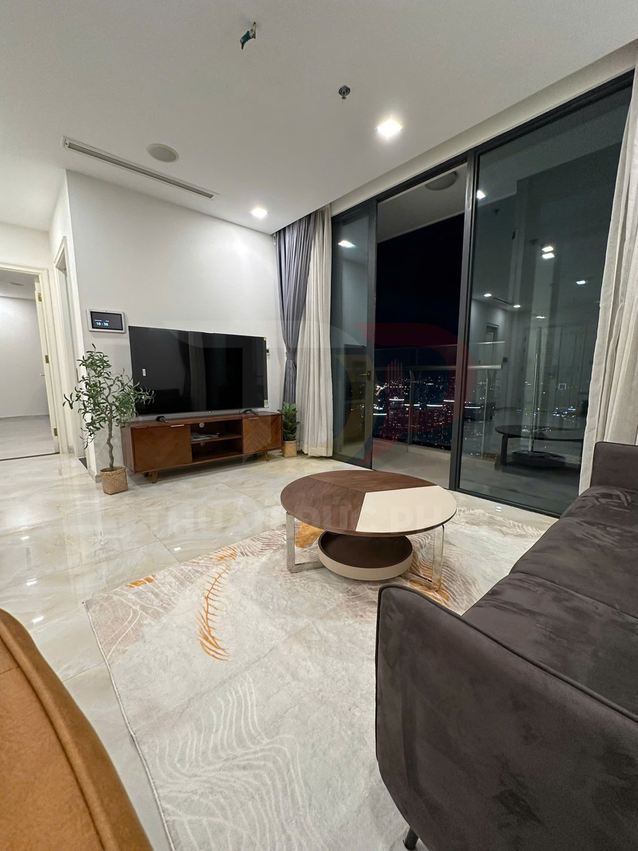 Bàn trà tròn hiện đại đặt trên thảm phòng khách cạnh sofa và kệ tivi gỗ, nhìn ra cửa kính ban công ban đêm