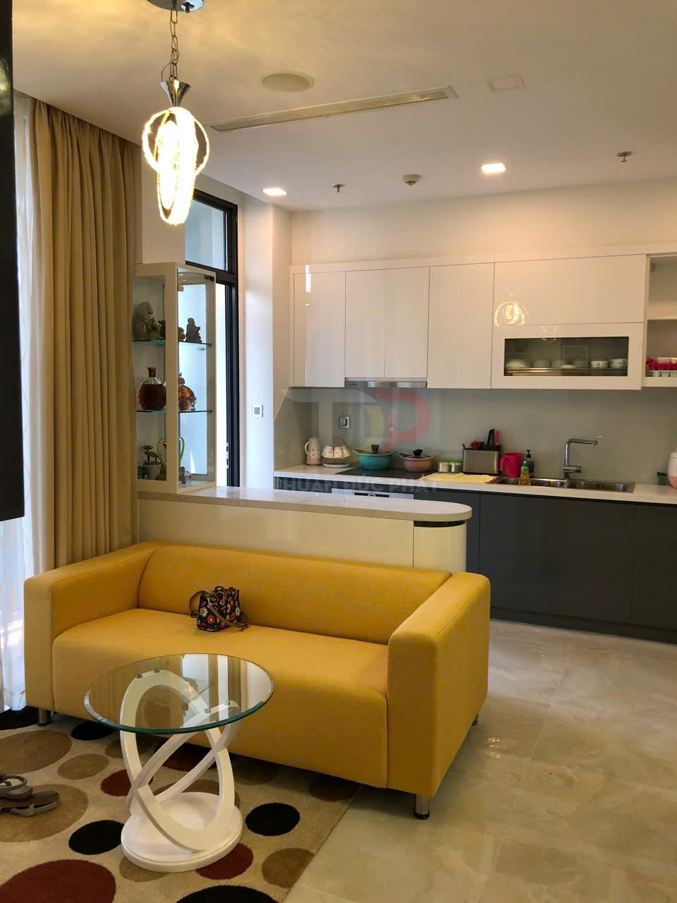 Sofa văng màu vàng đặt cạnh bàn trà kính tròn chân xoắn trong phòng khách liền bếp hiện đại