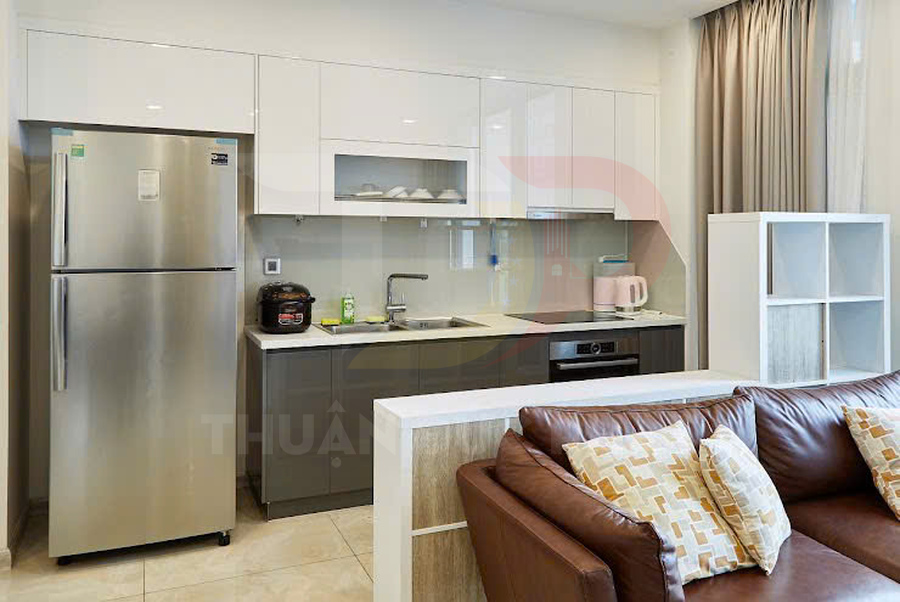 Bếp nhỏ hiện đại với tủ bếp trắng, tủ lạnh inox, bếp âm và sofa da nâu trong căn hộ tiện nghi