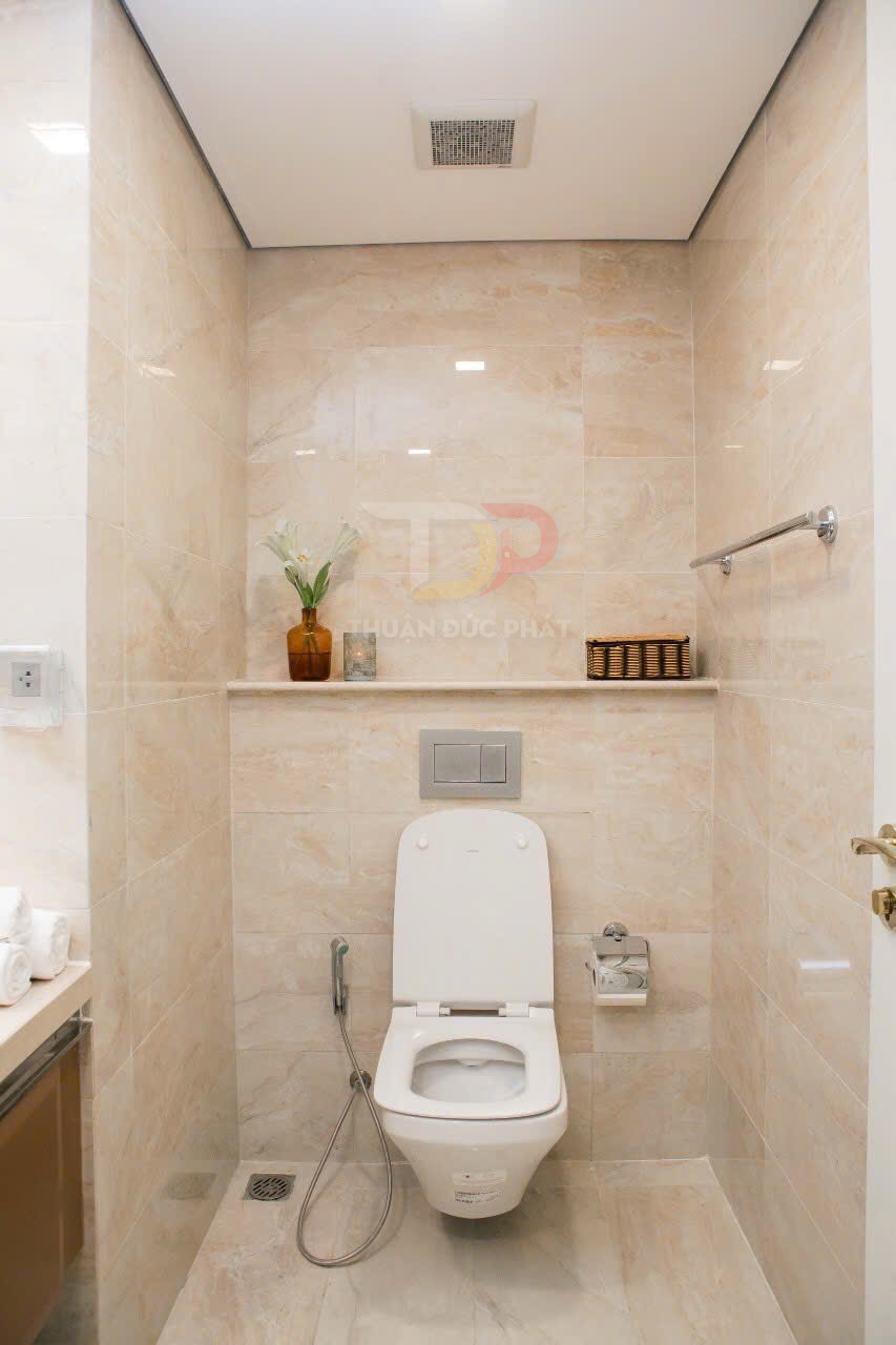 Bồn cầu sứ trắng hiện đại trong nhà vệ sinh ốp gạch sáng màu có vòi xịt và kệ trang trí