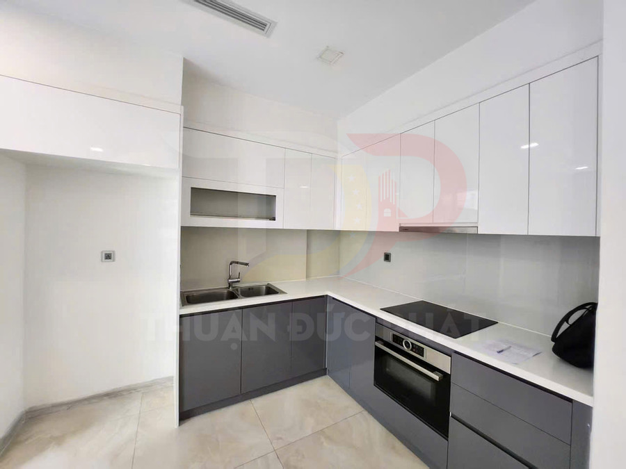 Gian bếp hiện đại với tủ bếp chữ L, bếp từ âm, lò nướng và chậu rửa inox trên nền trắng sáng