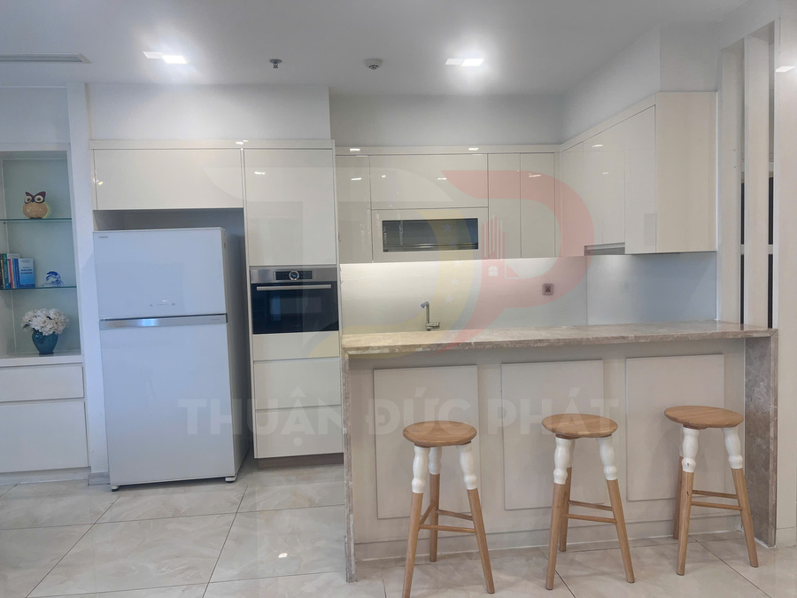 Bếp hiện đại với tủ bếp trắng, tủ lạnh, lò nướng âm tủ và quầy bar đá kèm ba ghế gỗ