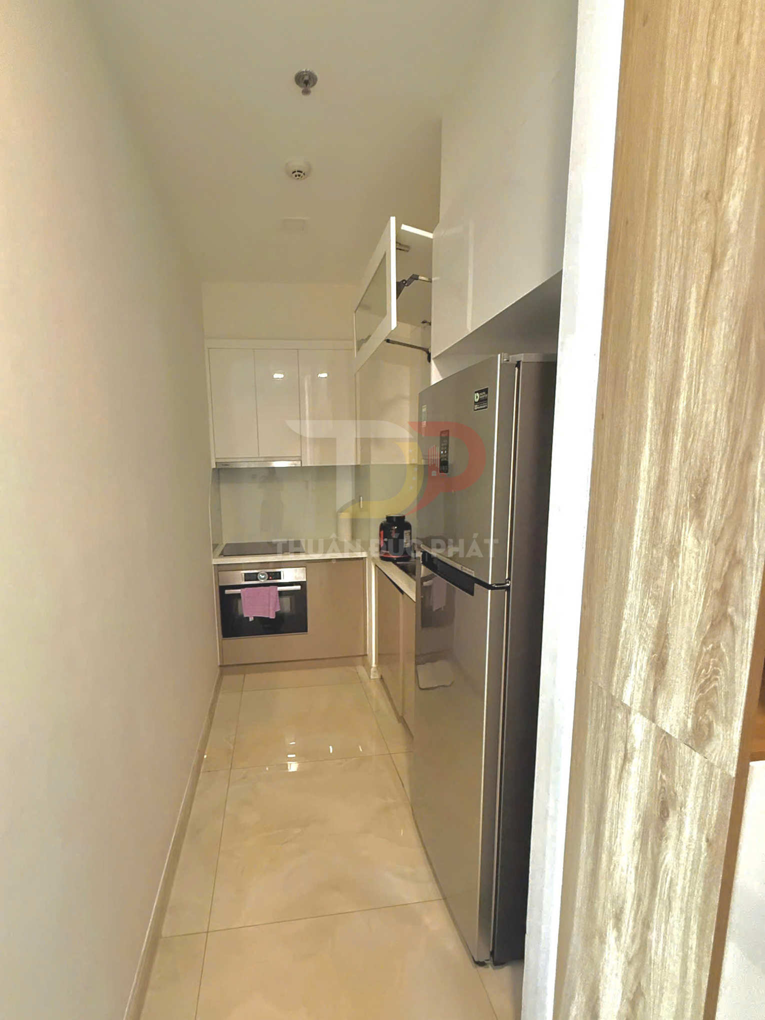 Gian bếp nhỏ hiện đại với tủ bếp trắng, bếp âm và tủ lạnh inox cạnh lối đi hẹp