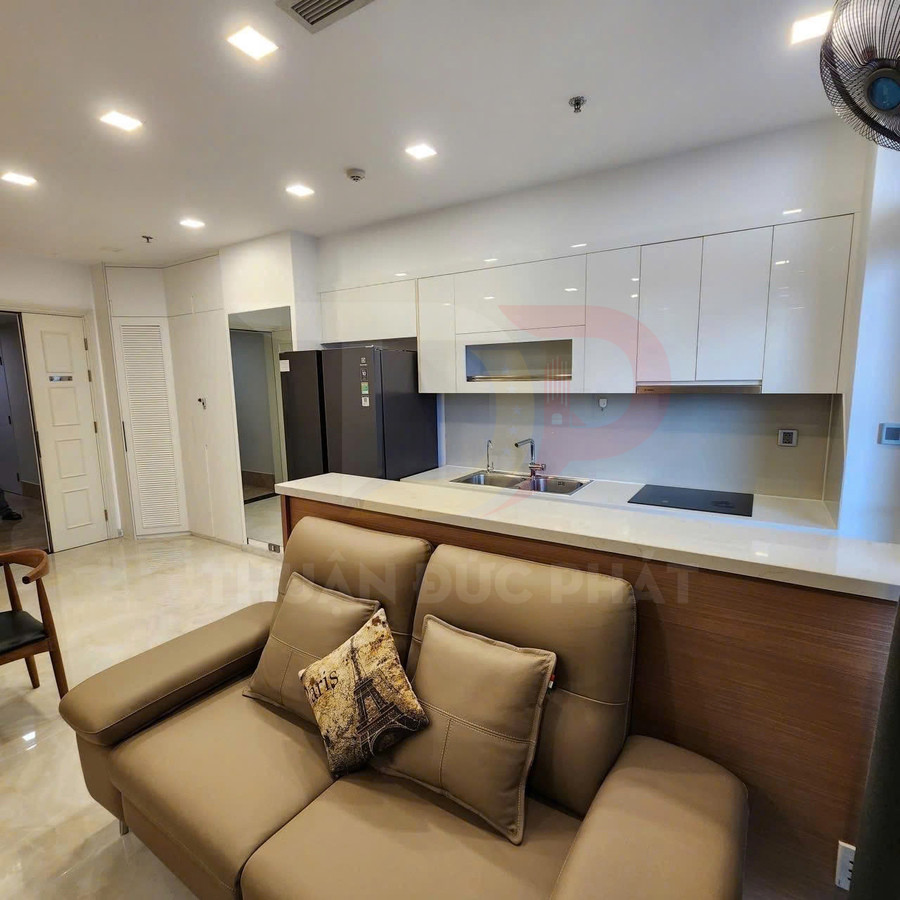 Sofa da màu nâu đặt trước bếp hiện đại với tủ bếp trắng, bồn rửa đôi và bếp từ âm tường