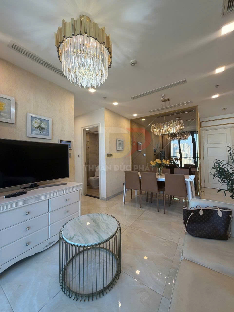 Phòng khách chung cư hiện đại với đèn chùm pha lê, bàn trà mặt kính và bàn ăn sáu ghế tông nâu sáng