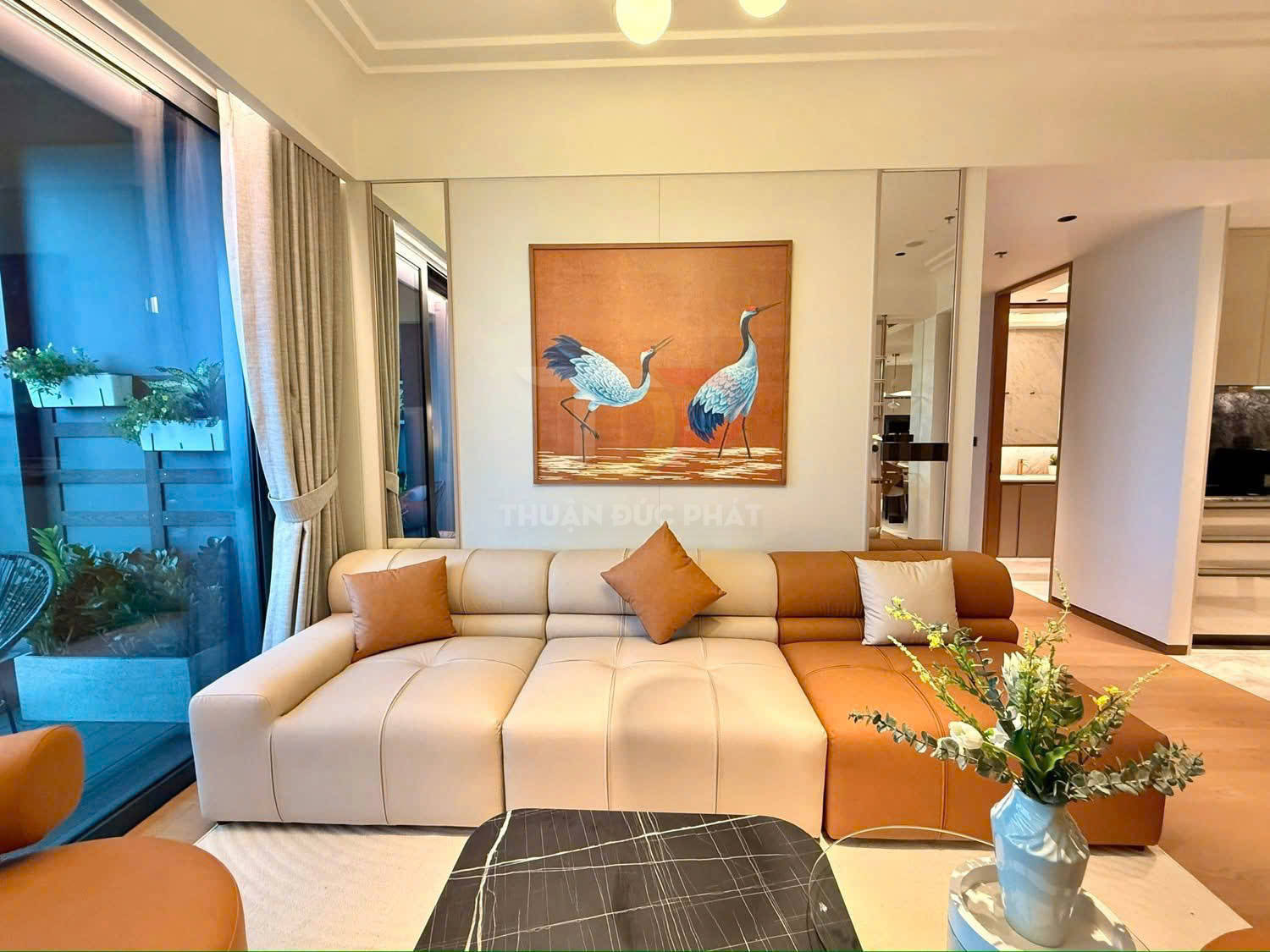 Sofa da chữ L màu kem nâu trong phòng khách hiện đại, bàn trà đá đen và tranh treo tường đôi chim hạc