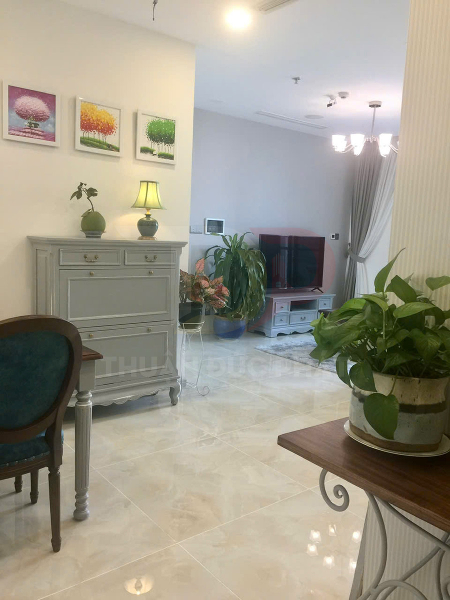 Phòng khách lát gạch bóng màu kem với tủ gỗ sơn xám, cây xanh trang trí và tranh treo tường nhiều màu