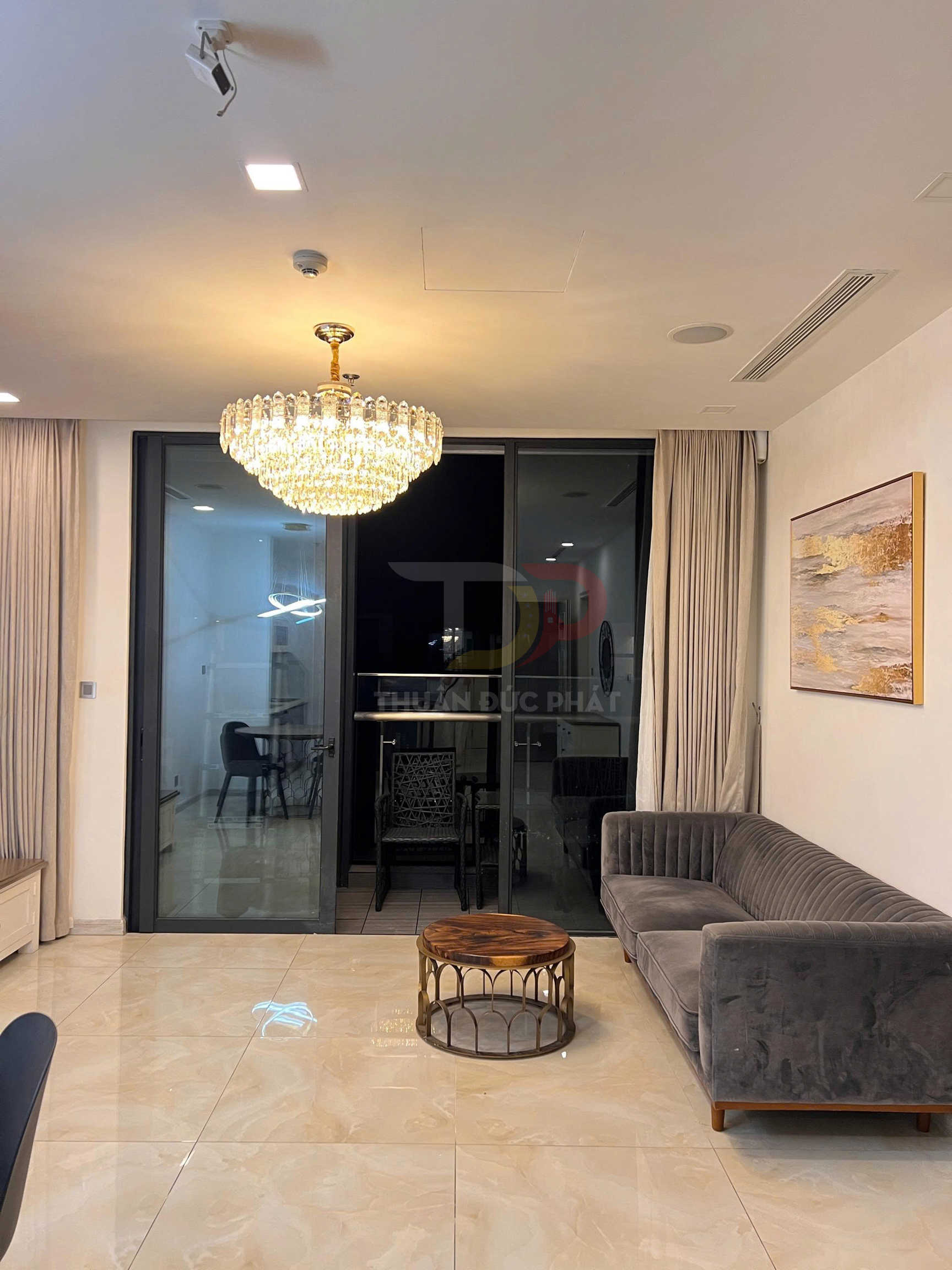 Đèn chùm pha lê trần phòng khách hiện đại với sofa nhung xám và bàn trà tròn mặt gỗ