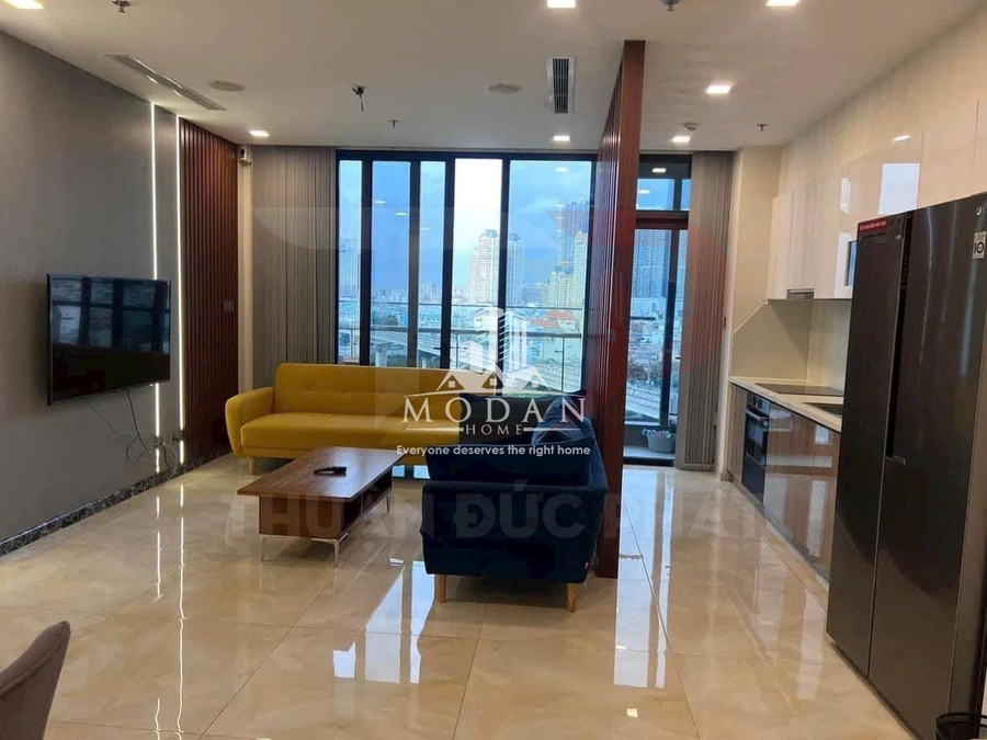 Phòng khách căn hộ hiện đại với sofa vàng, bàn gỗ, tivi treo tường và bếp liền kề nhìn ra cửa kính lớn