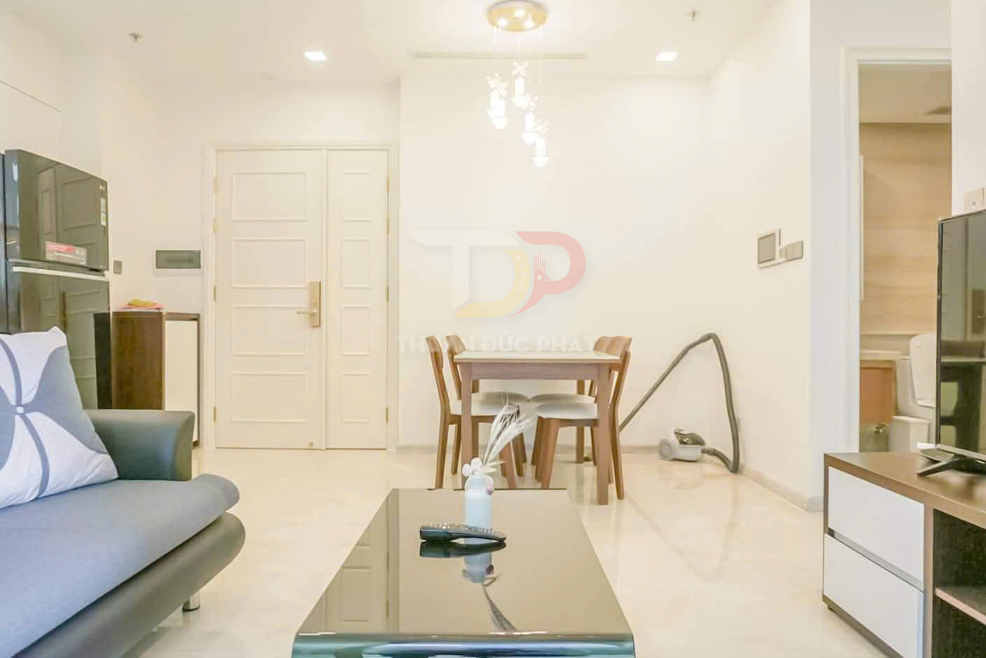 Phòng khách căn hộ hiện đại với sofa, bàn trà kính, bàn ăn gỗ và máy hút bụi đặt sát tường