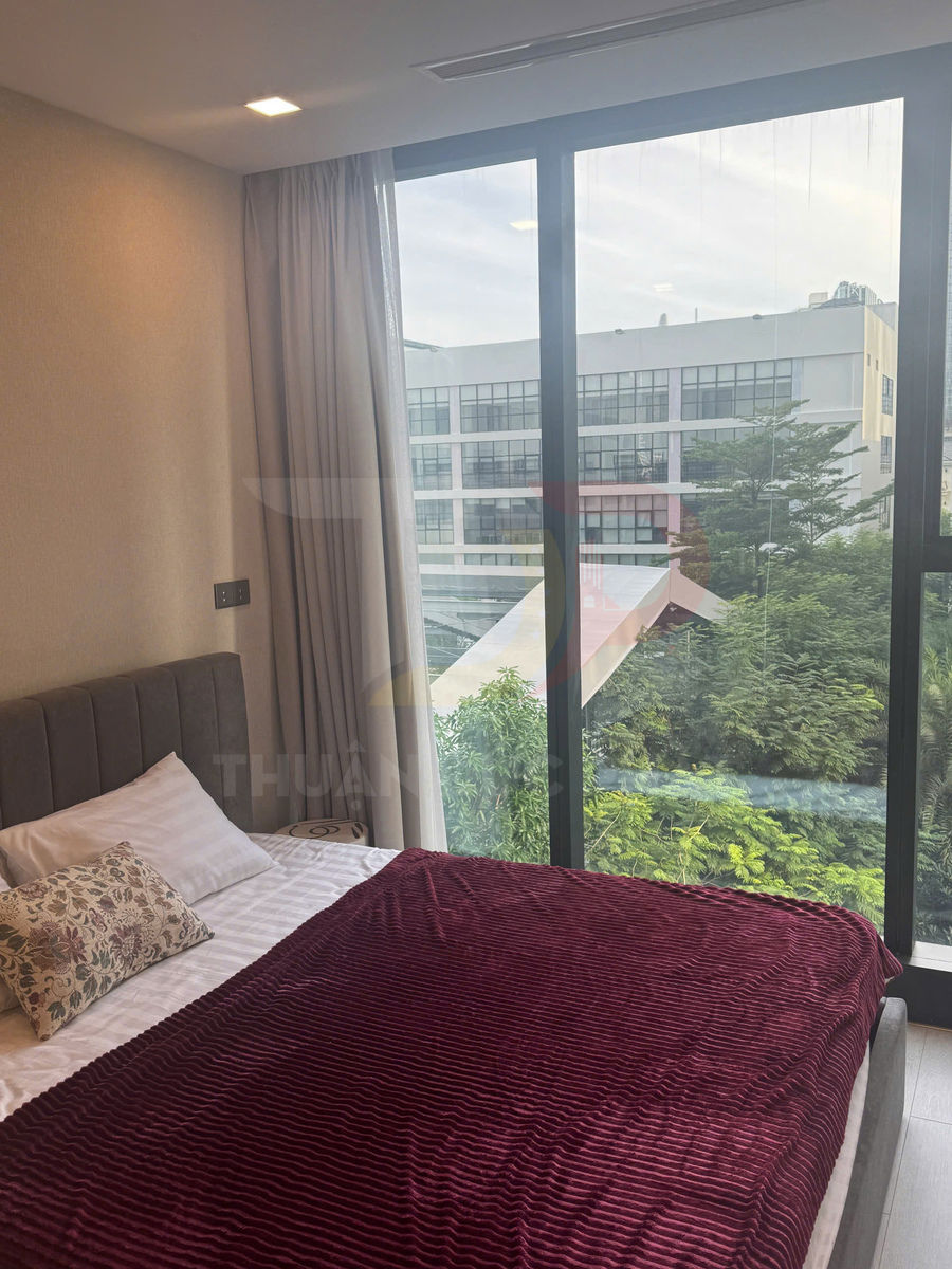 Phòng ngủ hiện đại với giường đôi chăn nhung đỏ cạnh cửa kính lớn nhìn ra cây xanh và tòa nhà văn phòng