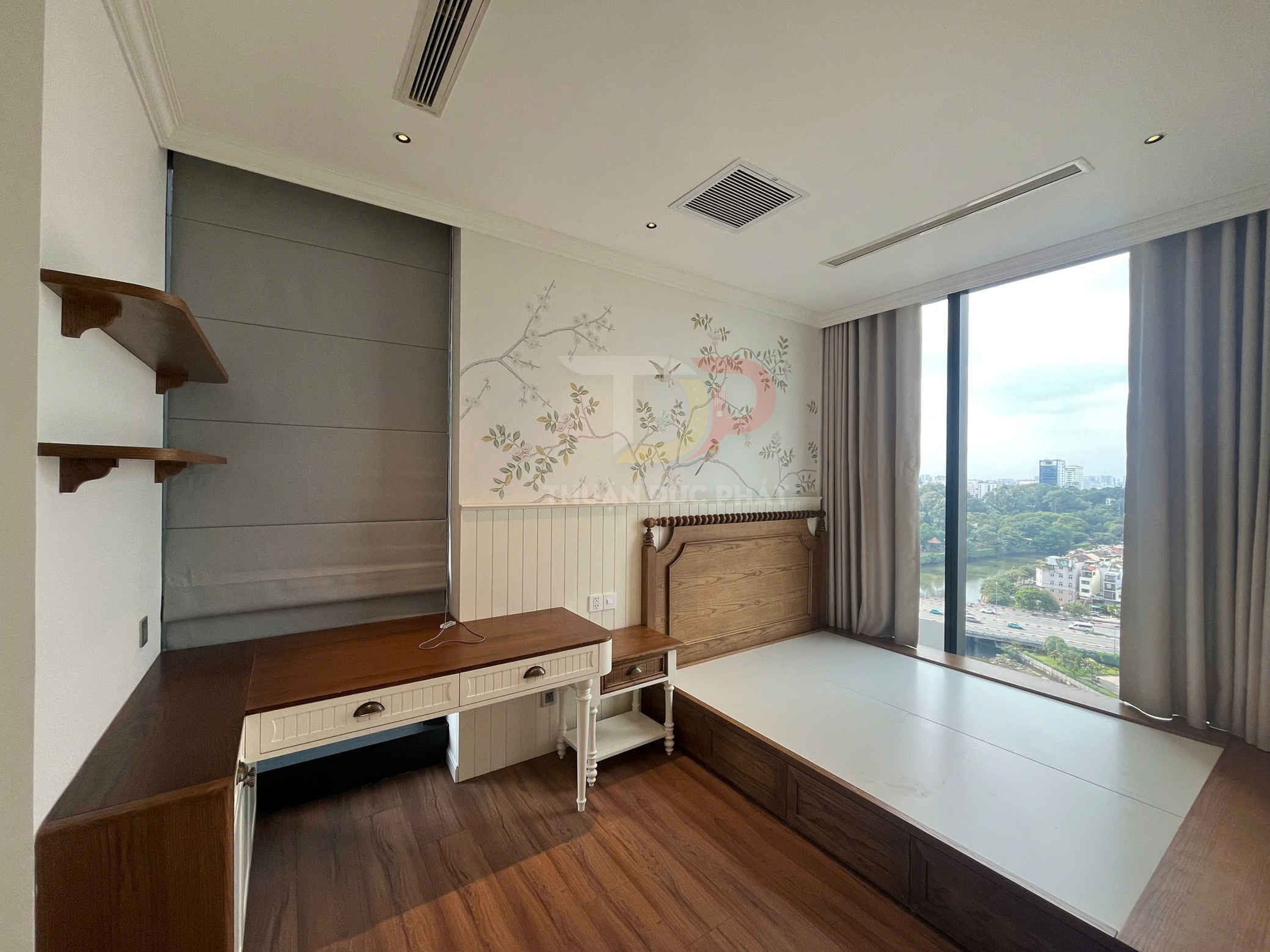 Phòng ngủ nhỏ với giường gỗ liền khối, bàn làm việc gỗ và cửa sổ kính lớn nhìn ra thành phố