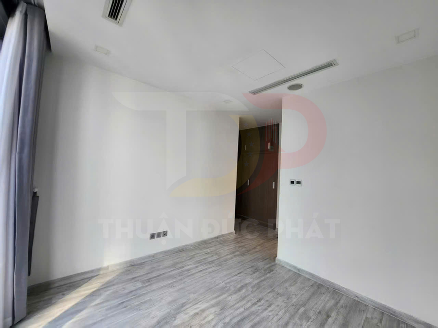 Phòng ngủ trống lát sàn gỗ, tường trắng, cửa sổ rèm xám và tủ âm tường gỗ hiện đại