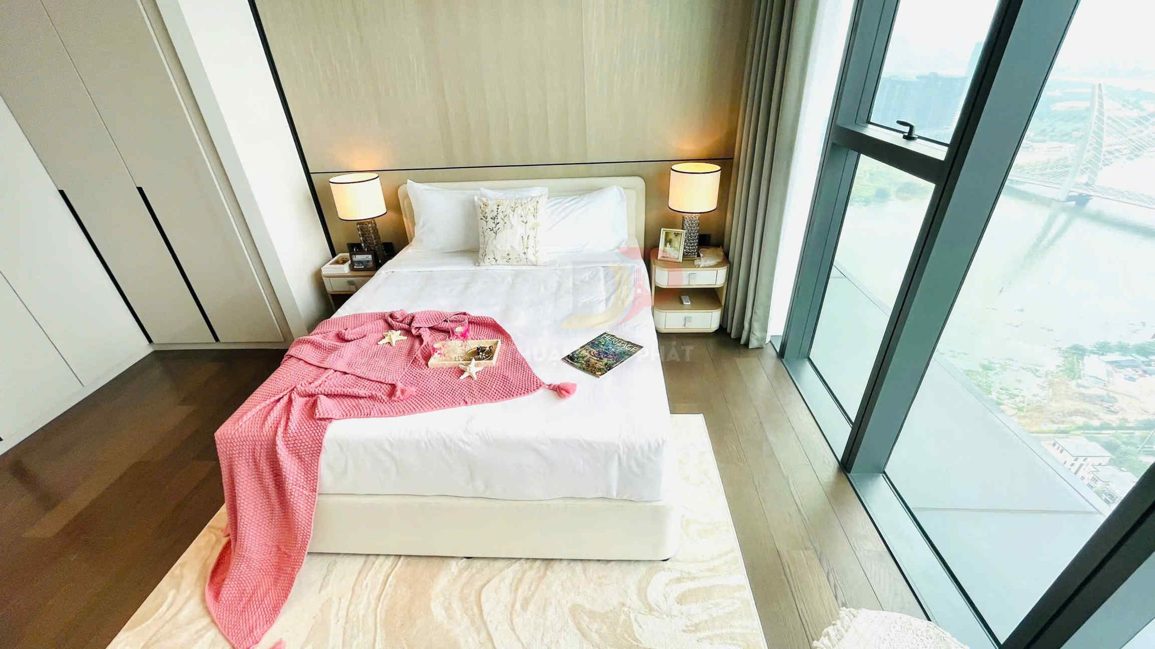 Giường ngủ đôi trải ga trắng với chăn len hồng, bàn đầu giường và cửa sổ kính lớn nhìn ra thành phố