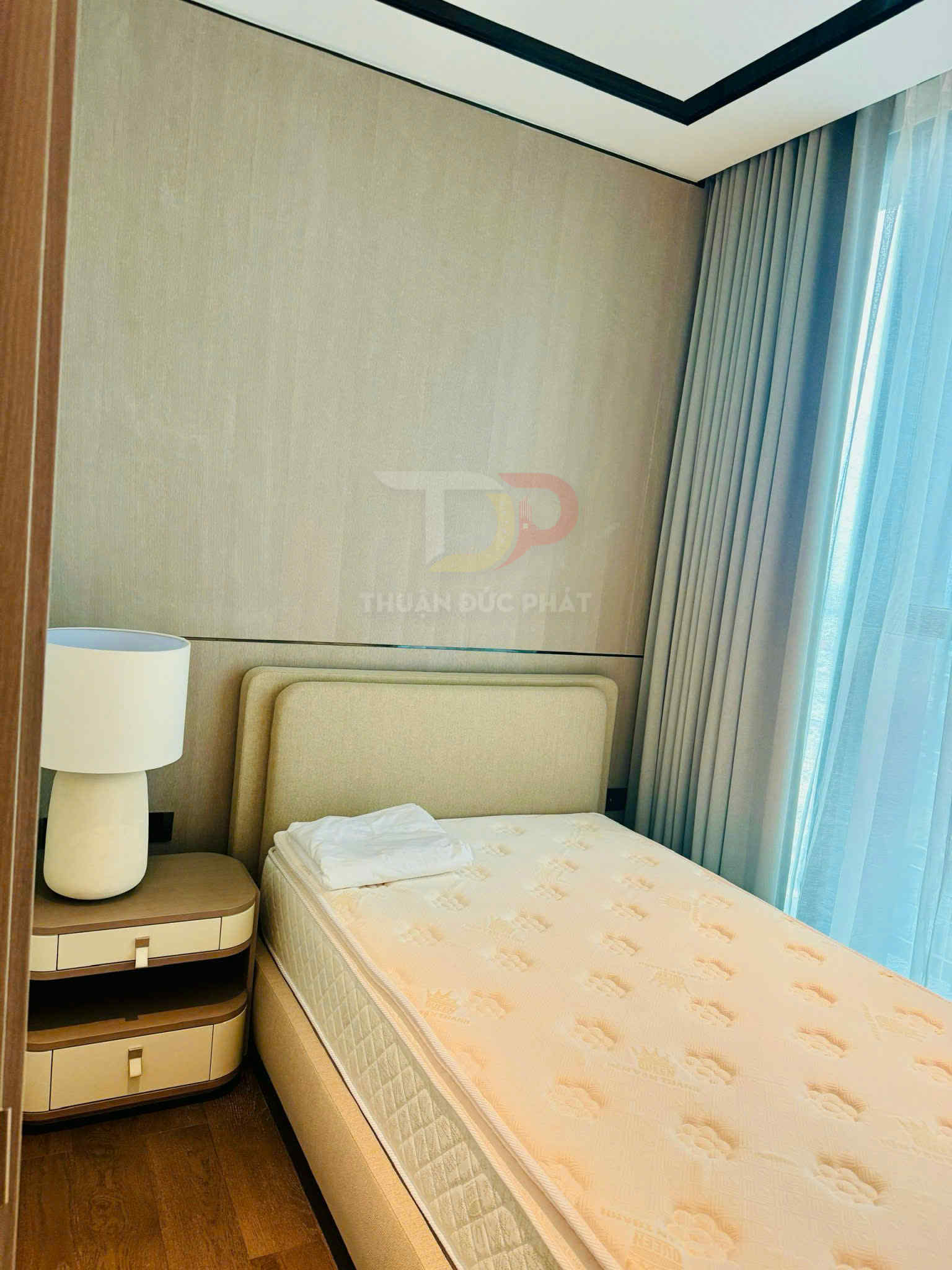 Giường ngủ đơn hiện đại với nệm màu kem, tủ đầu giường và đèn trang trí cạnh cửa sổ có rèm sáng màu