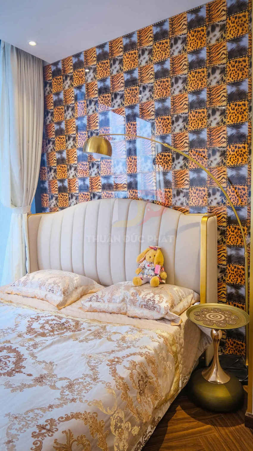 Giường ngủ bọc nệm màu kem với chăn ga hoa văn vàng, tường sau dán giấy họa tiết da thú nổi bật