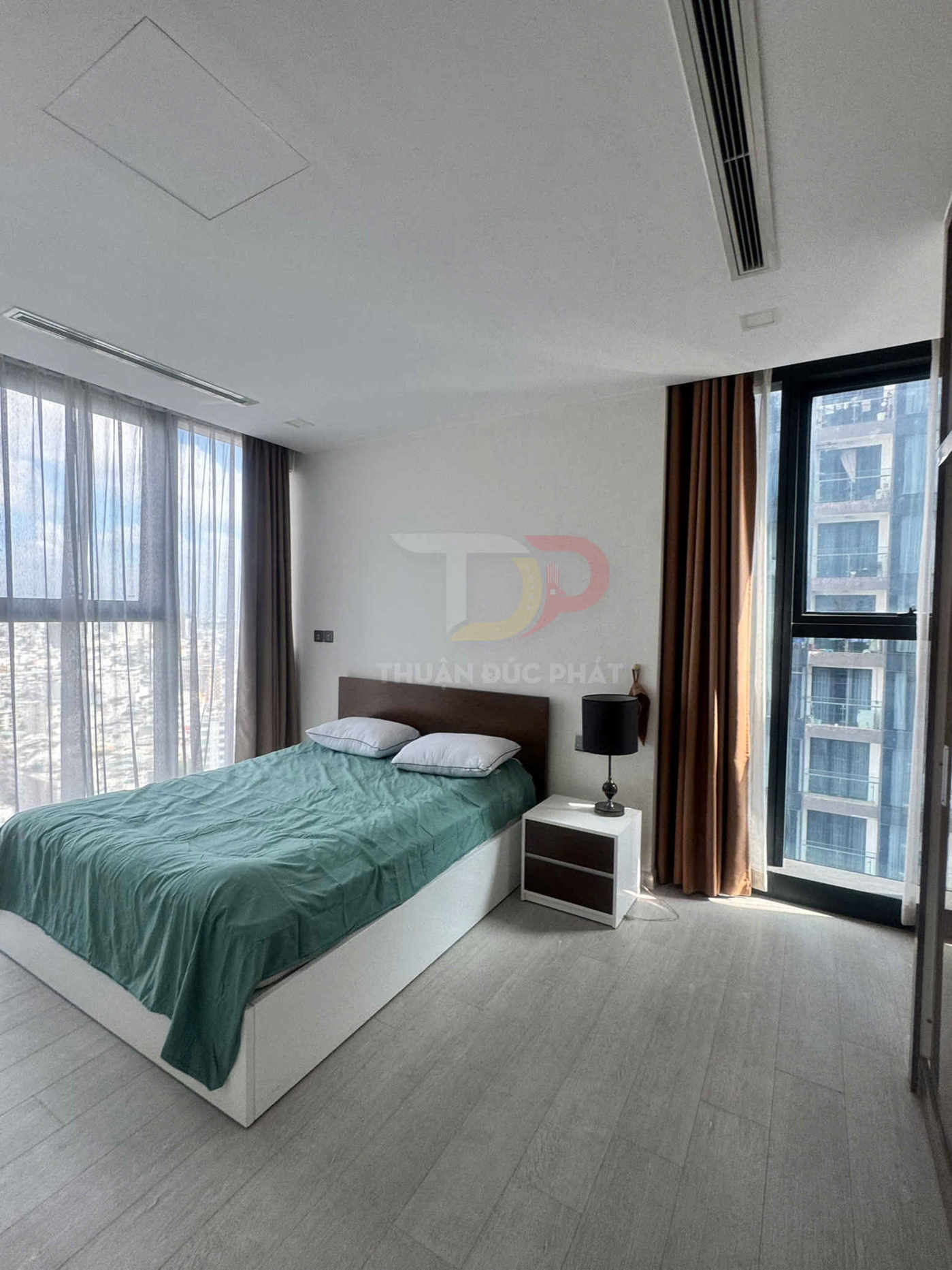 Phòng ngủ hiện đại với giường đôi ga xanh, tủ đầu giường trắng nâu và cửa sổ kính lớn nhiều ánh sáng