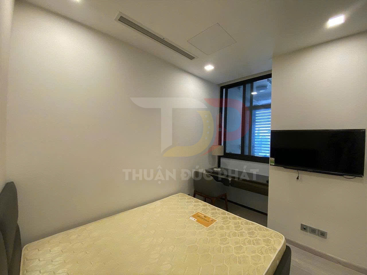 Phòng ngủ nhỏ với nệm giường mới, bàn làm việc cạnh cửa sổ kính và tivi treo tường