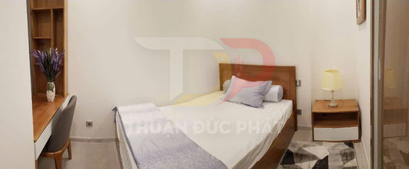 Phòng ngủ nhỏ với giường gỗ, ga trắng, bàn làm việc và tủ đầu giường có đèn ngủ