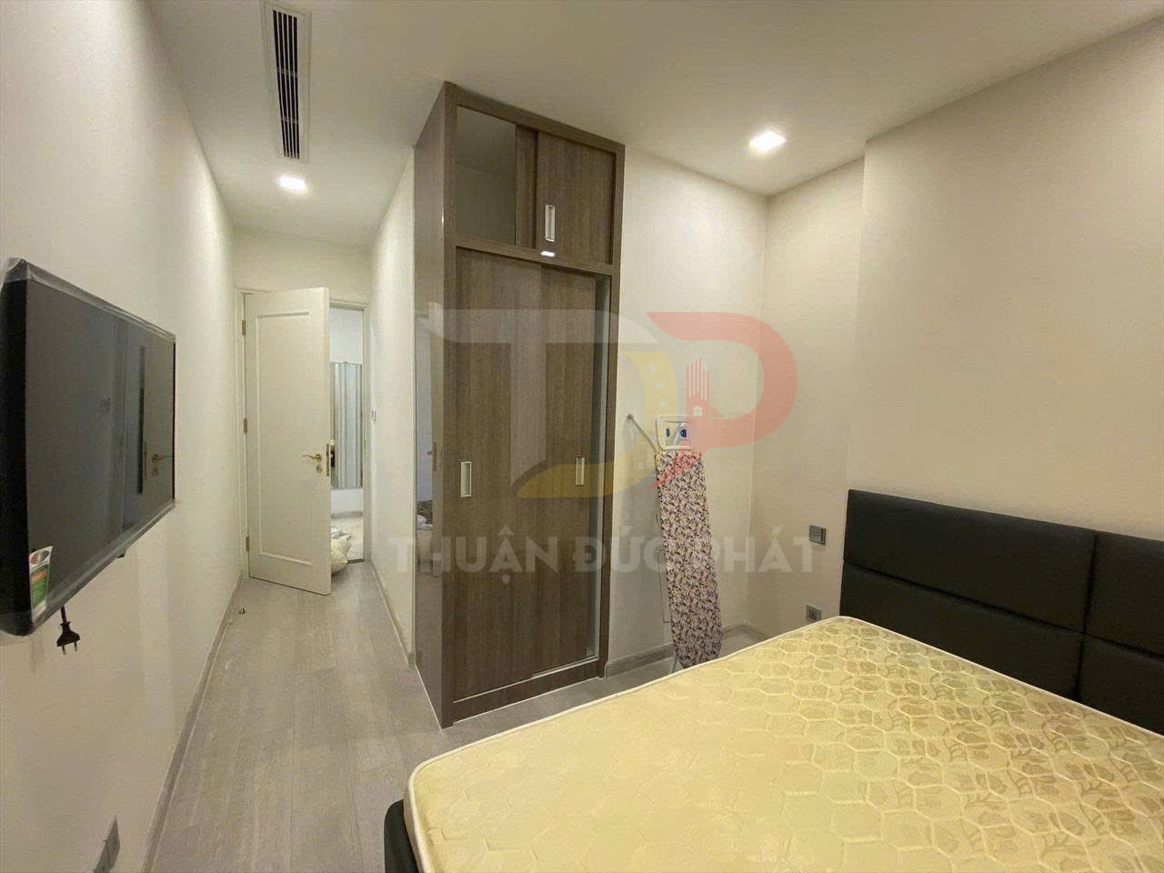 Phòng ngủ nhỏ hiện đại với giường nệm, tủ quần áo gỗ, bàn ủi đứng và tivi treo tường