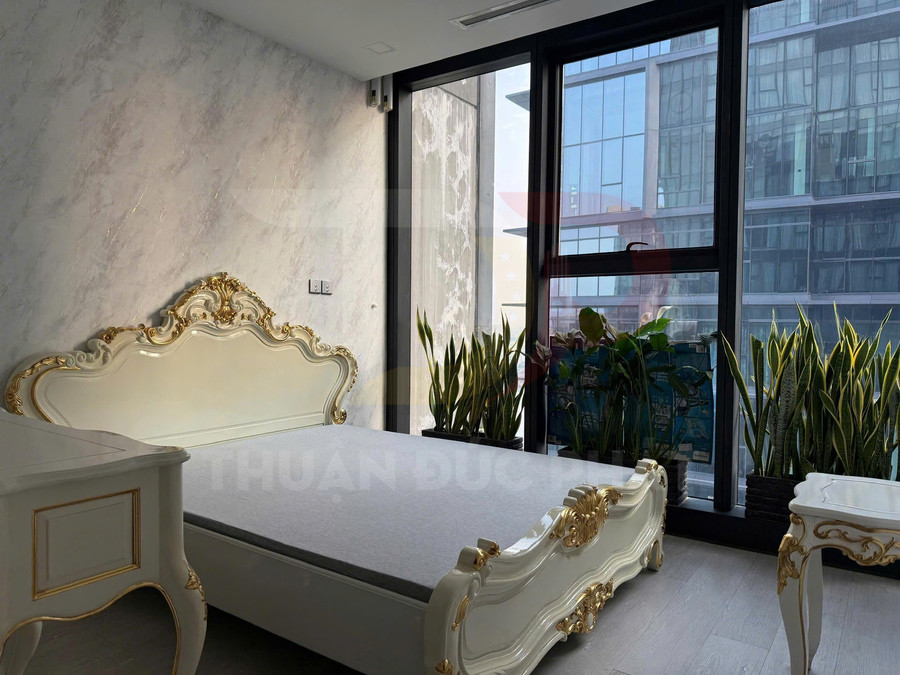 Giường ngủ tân cổ điển màu trắng viền vàng cạnh cửa sổ kính lớn và chậu cây xanh trong phòng ngủ hiện đại