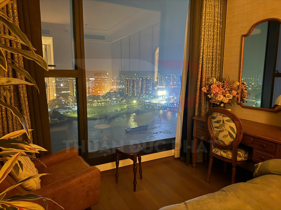 Phòng ngủ cao cấp với bàn trang điểm gỗ, ghế hoa văn và cửa sổ lớn nhìn ra sông và thành phố về đêm